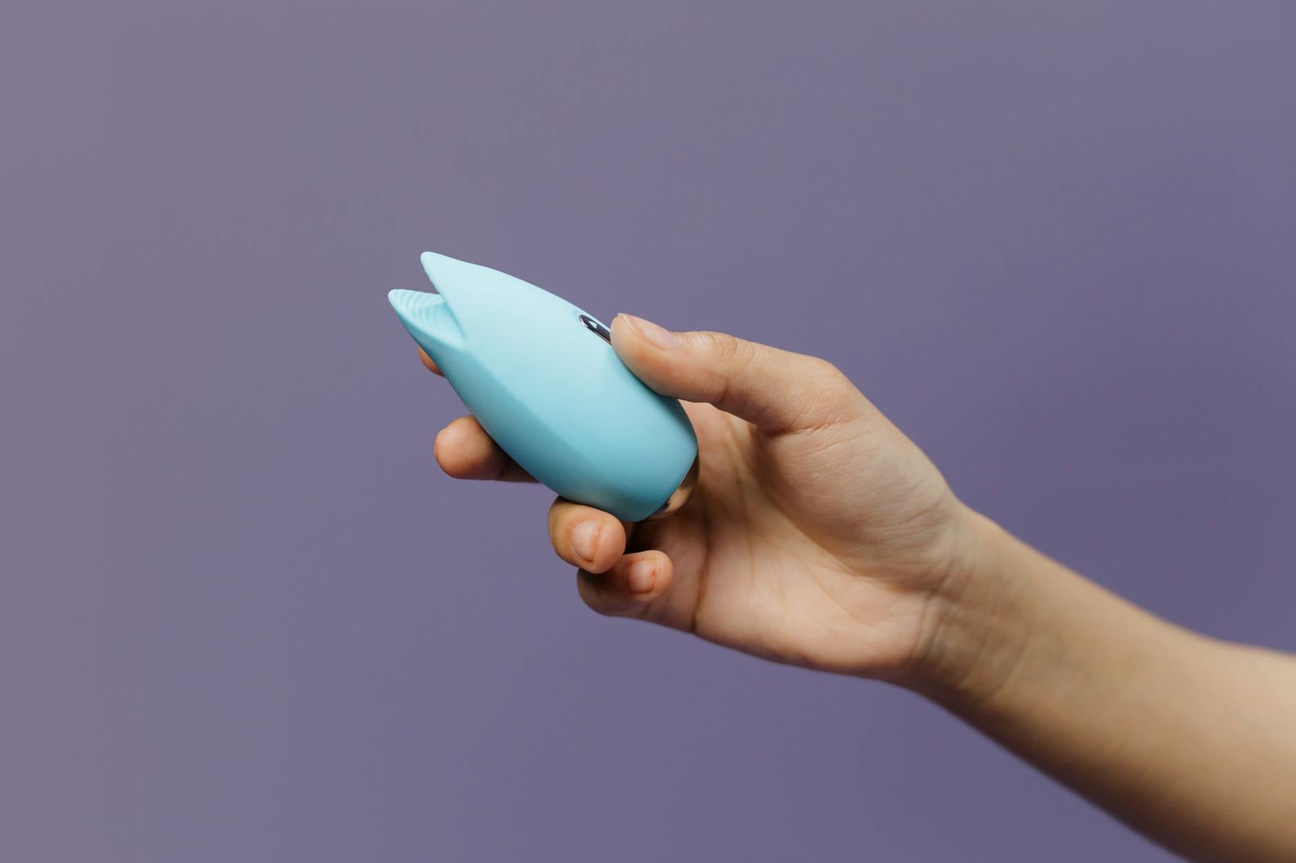 A hand holding a blue silicone vibrator against a purple background, representing modern clitoral stimulation tools